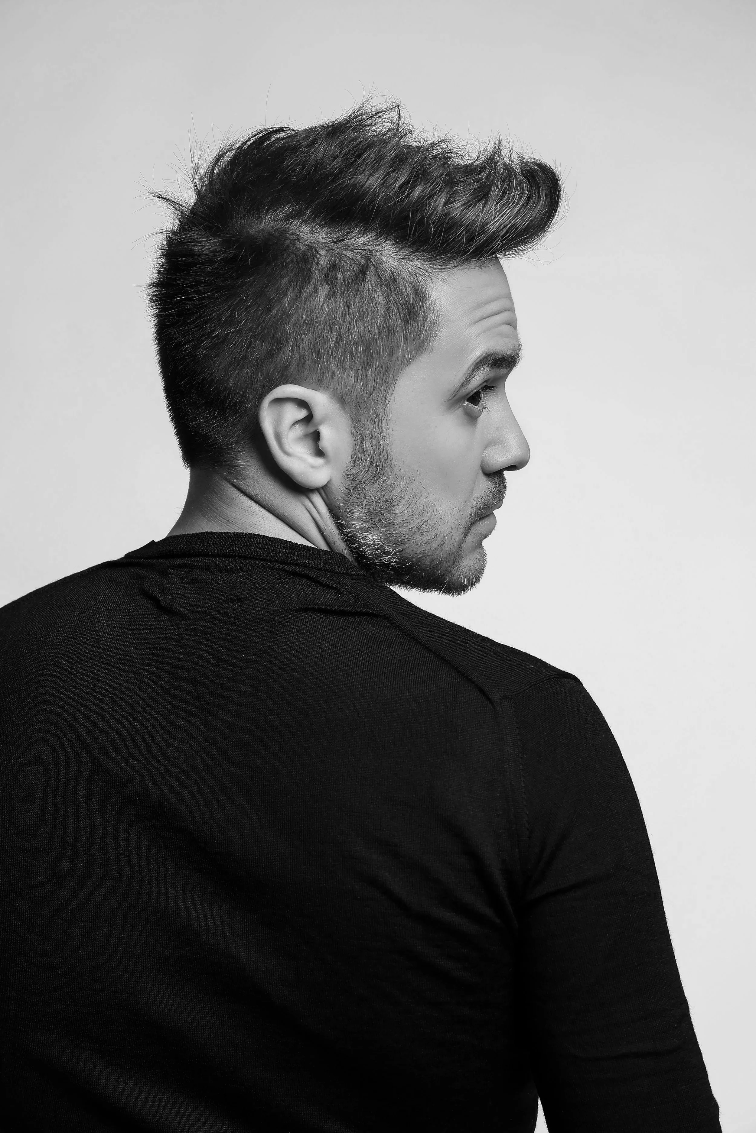 Portrait d'un homme vu de côté, portant un t-shirt noir, avec une coiffure courte sur les côtés et cheveux plus longs sur le dessus, cheveux en bataille, barbe légère, fond clair.